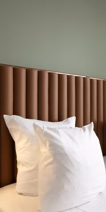 A modern hotel room with a double bed featuring a padded brown headboard against a green wall. Two white pillows are stacked on each side. Black wall-mounted reading lights are attached to the headboard. Cream curtains are partially visible.