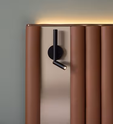 Close-up of a modern bed headboard with vertical brown leather panels against a green wall. A black, minimalist wall lamp is mounted above the headboard, and warm light glows softly from the top. A white pillow is partially visible.