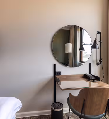 A minimalist hotel room corner features a round mirror above a wooden desk with a chair. A wall-mounted lamp and a telephone are on the desk. Nearby, a wall-mounted TV and a portion of a bed with white sheets are visible. A window with beige curtains is on the right.