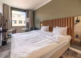 Modern hotel room with a large bed, white linens, and a padded headboard. A desk with a chair is in the left corner, and a small round table with an armchair is near the window. Neutral tones and natural light create a cozy atmosphere.