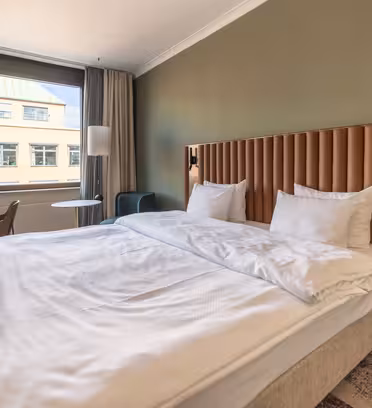 Modern hotel room with a large bed, white linens, and a padded headboard. A desk with a chair is in the left corner, and a small round table with an armchair is near the window. Neutral tones and natural light create a cozy atmosphere.