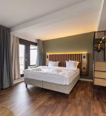 A modern hotel room with a neatly made double bed with white linens. The room has wooden floors, a TV on the wall, a tall lamp and large windows with curtains. There is a small round table and a stylish bookcase next to the bed.