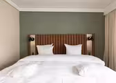 A neatly made bed with white sheets and two pillows against a green accent wall. The headboard is brown with vertical tufting. Two wall-mounted reading lights are on either side. Beige curtains hang on the right.