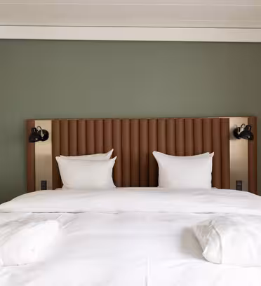 A neatly made bed with white sheets and two pillows against a green accent wall. The headboard is brown with vertical tufting. Two wall-mounted reading lights are on either side. Beige curtains hang on the right.