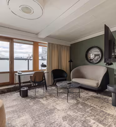 A modern hotel room features a plush sofa, a round mirror, and a large wall-mounted TV. A small table with chairs and a lamp sit by the window. Neutral tones and a patterned carpet enhance the cozy atmosphere. Large windows show a cloudy sky outside.