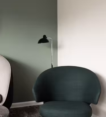 A modern interior features a beige armchair and a dark green chair. A black, round mirror hangs on a green wall, and a black floor lamp stands next to it. A white wall contrasts the corner of the room.