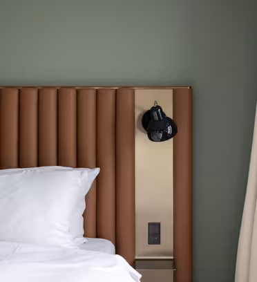 A modern hotel room with a bed with white pillows and sheets. The headboard is brown and vertically grooved. A black wall lamp is mounted on the headboard, next to a beige wall and beige curtains. The wall is painted green.