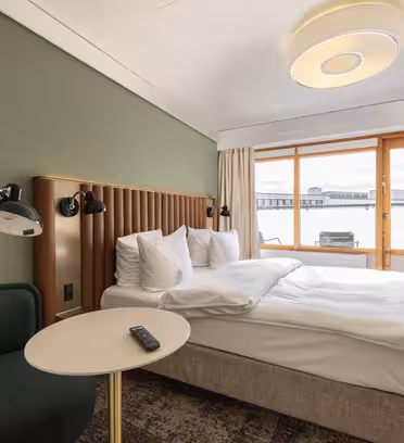A modern hotel room features a neatly made double bed with white linens, a green accent wall, and a padded headboard. Theres a small round table with a chair beside it. A large window offers a view of a cityscape with industrial buildings.