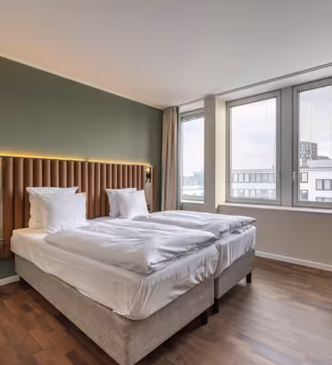 A hotel room with two neatly made beds against a green accent wall. The room has large windows overlooking the city, a small round table, and a dark green armchair. Natural light fills the minimalist space, highlighting the wooden floor.
