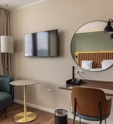 Modern hotel room featuring a round mirror above a desk with a chair and phone. A wall-mounted TV is beside a window with light curtains. A green armchair and small round table are in the corner, with a floor lamp nearby.