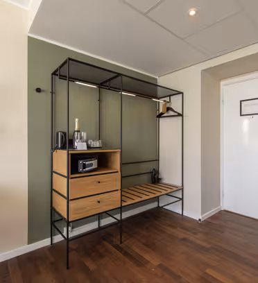 Modern hotel room entryway with a minimalist design. Features a wall-mounted lamp, circular mirror, and a wooden shelving unit with a mini fridge, electric kettle, and champagne bottle. The floor is wooden, and the door is white with a digital lock.