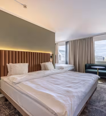 Hotel room with a large bed, an additional single bed, and modern decor. Features a green accent wall, bedside lamps, a small seating area with a green sofa and round table, large windows with light curtains, and a city view.