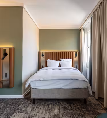 A modern hotel room with two single beds, featuring upholstered headboards and soft white bedding. The walls are light green and cream, with curtains drawn to one side. The floor is carpeted, and a small wall lamp is above each bed.