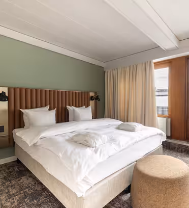 A modern bedroom with a large bed featuring white bedding and pillows. Theres a padded headboard, green accent wall, two wall-mounted lights, a beige ottoman, and large windows with curtains leading to a small balcony.