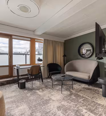 Modern hotel room with a large window, neutral tones, and minimal decor. Includes a gray sofa, black armchairs, round coffee table, rug, wall-mounted TV, and circular mirror. Curtains partially cover the window revealing an overcast sky outside.
