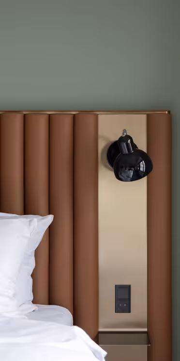 A modern bedroom features a bed with white pillows and a white sheet. The bed has a brown, vertically ribbed headboard, and a black wall-mounted lamp is attached to a beige panel. Light-colored curtains hang on the right.