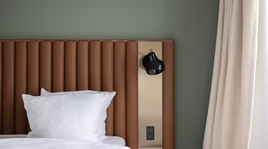 A modern bedroom features a bed with white pillows and a white sheet. The bed has a brown, vertically ribbed headboard, and a black wall-mounted lamp is attached to a beige panel. Light-colored curtains hang on the right.