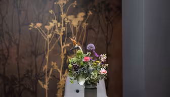 A vase of colorful flowers, including pink, purple, and green blooms, sits on a chair in front of a dark wall with a golden floral pattern. The background creates a dramatic contrast with the vibrant flowers in the foreground.