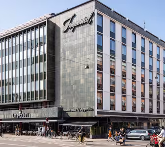 A modern multi-story building with a grid of windows and a dark exterior, featuring the word Imperial on the facade. The ground floor has a restaurant with outdoor seating. Cyclists and pedestrians are visible on the street.