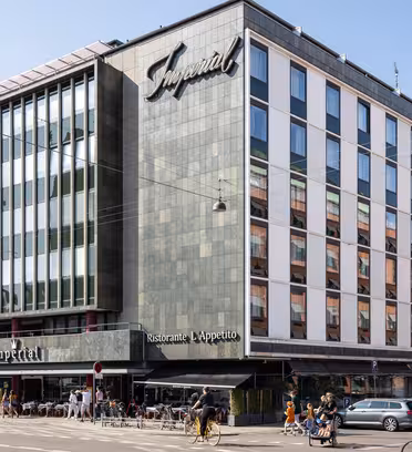 A modern multi-story building with a grid of windows and a dark exterior, featuring the word Imperial on the facade. The ground floor has a restaurant with outdoor seating. Cyclists and pedestrians are visible on the street.