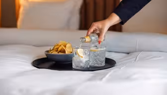A hand pours a clear beverage from a small bottle into a glass with ice and lemon, next to another similar glass and a bowl of chips, all on a tray on a neatly made bed.
