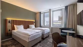 Modern hotel room with two single beds, a padded headboard, green accent wall, large windows with city view, a small table with chairs, and a wall-mounted TV. Neutral colors and contemporary decor.
