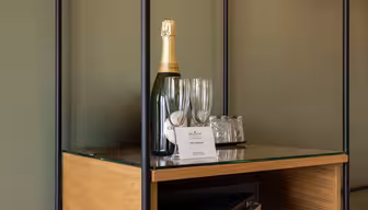 A bottle of sparkling wine, two empty champagne flutes, and two glasses on a tray sit on a glass shelf with a wooden base. A small card is placed in front of the tray.