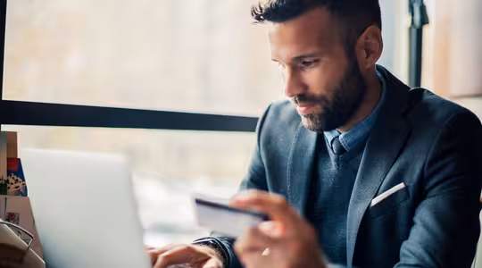 A man sits at a table by the window, concentrating on his laptop. Hes wearing a gray suit and holding a credit card, suggesting online shopping or a financial transaction. The background is slightly blurred, emphasizing a bright, natural atmosphere.