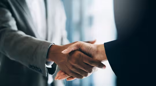 Close-up of a handshake between two individuals, one in a gray suit and the other in a black suit. The background is blurred, emphasizing the gesture of agreement or partnership.