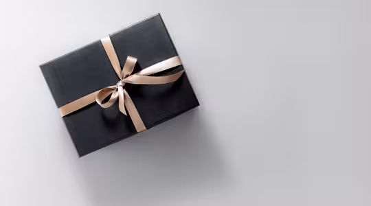 A black gift box with a shiny beige ribbon tied in a bow is positioned on the left side of a light gray background.