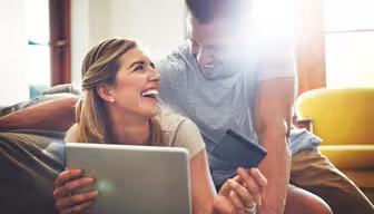 A smiling couple sits together on a couch in a bright room. The woman holds a tablet and a credit card, while the man leans over her shoulder, both looking at each other joyfully. A yellow chair and sunlight are visible in the background.