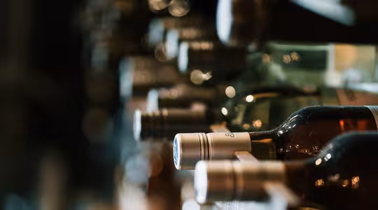 Close-up of a wine rack holding several bottles of wine, each with a metal screw cap. The bottles are arranged horizontally, with various colors visible through the glass. The background is softly blurred, emphasizing the caps in the foreground.