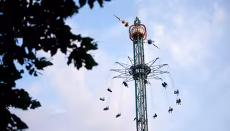 A tall swing ride at an amusement park with people seated in swings hanging from a rotating circular platform. The ride is set against a blue sky, partially visible through tree leaves in the foreground.