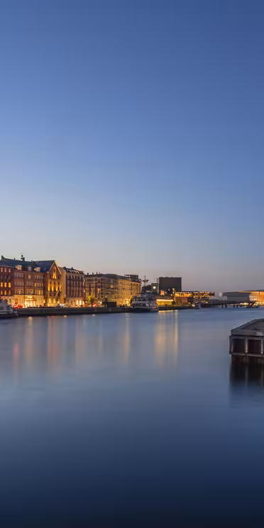 Waterfront view of a city at dusk, showcasing calm waters and buildings with illuminated windows. The sky is a gradient from light to deep blue, and the scene reflects on the water, creating a peaceful and serene atmosphere.