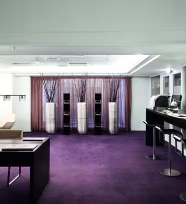 Modern office space with purple carpet, featuring a long high desk with computers and stools, a separate desk with chairs, and three large vases filled with branches near a sheer curtained window. Neutral color scheme and sleek design elements.