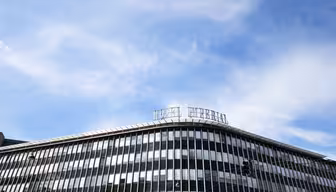 A modern building with large horizontal windows is set against a blue sky with wispy clouds. A rooftop sign is visible, and a traffic light can be seen in the foreground.
