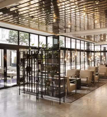 A modern, stylish cafe with glass walls and a reflective ceiling. Indoor plants are placed on shelves, and there are beige chairs around tables. The space is bright, with large windows and elegant decor.