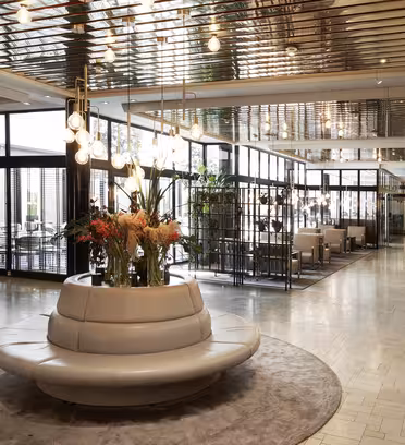 Hotel lobby with a modern design featuring a circular gray seating area with flowers in the center. Overhead, globe lights hang from the mirrored ceiling. Glass walls enclose conference rooms, and a sign points to various amenities.