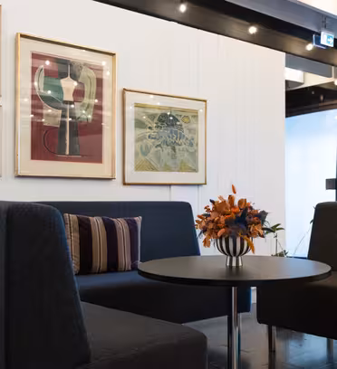 Modern office waiting area with two dark gray chairs and a round black table topped with a vase of orange flowers. The white wall behind has framed abstract art. Ceiling lights illuminate the space.