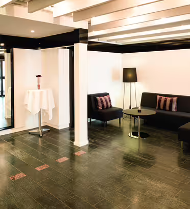 A modern lounge area with dark tiled flooring and white walls. It features two black sofas with striped cushions, two black floor lamps, and a round coffee table. A large mirror and a tall cocktail table with a white cloth are nearby.