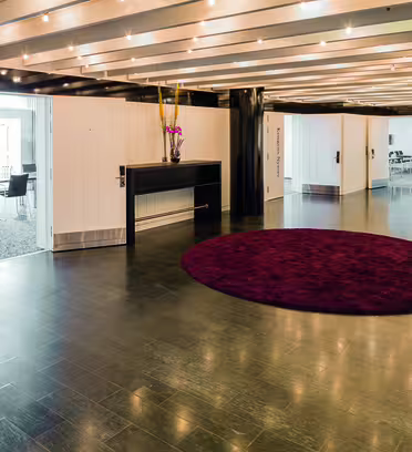 Spacious modern lobby with white walls and ceiling lights. A round burgundy rug is in the center, with small tables and floral arrangements along the sides. Open doors lead to rooms labeled Amagertorv and Nytorv.