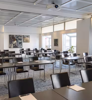 Empty conference room with rows of tables and black chairs facing a projector screen. Notepads and pens are on the tables. Large windows on the side walls let in natural light. Artwork hangs on one wall, and plants stand in corners.