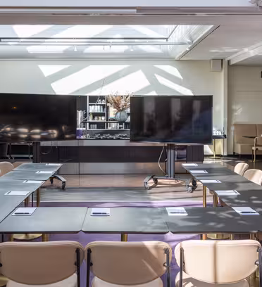 A modern conference room with U-shaped arrangement of tables and beige chairs. Two large screens are at the front. Natural light streams in through a skylight, illuminating the setting. Notepads are placed on each table.