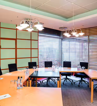 A modern conference room features a large U-shaped wooden table surrounded by black chairs. Water bottles and notepads are placed on the table. The room has a ceiling light fixture and is enclosed by frosted glass and blinds.