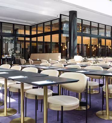A modern cafe interior with several empty black tables, each paired with cream-colored chairs. The space is well-lit with hanging lights, large windows, and a purple carpet. Notepads are placed on the tables, suggesting a meeting setup.
