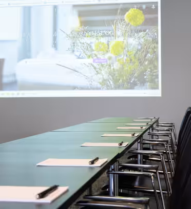 A conference room with a long table and chairs facing a screen displaying a projection of a website with flowers and blurred text. Notepads and pens are arranged neatly on the table.