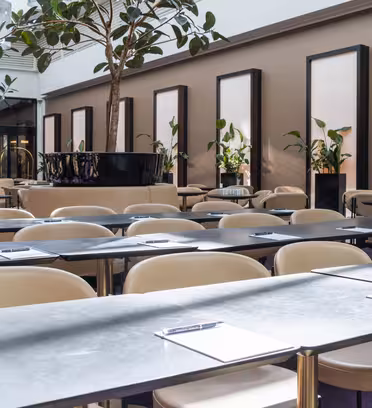 A modern conference room with rows of tables and beige chairs. Each table has a clipboard with paper and a pen. Tall plants in black pots are placed throughout the bright, spacious room with framed art and natural light from large windows.
