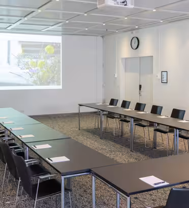 Conference room with U-shaped table setup, facing a large screen displaying a projected image. Black chairs are neatly arranged around the tables, and notepads are placed at each seat. Theres a clock and a closed door on the back wall.