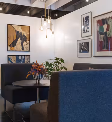 A modern lounge area with two dark gray sofas facing a round table, adorned with a colorful flower arrangement. Artworks decorate the white walls. Hanging pendant lights add a warm glow. Plants are placed in the corners of the room.