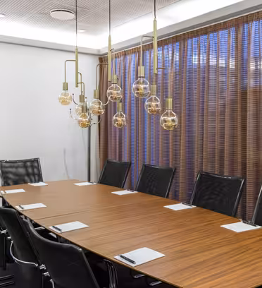 A modern conference room with a long wooden table surrounded by black leather chairs. Pendant lights with exposed bulbs hang above. The room has a clean, minimalist design, and a large window with sheer brown curtains.